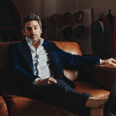 Mark Ghanime wearing a blue suit and posing while sitting on a couch.